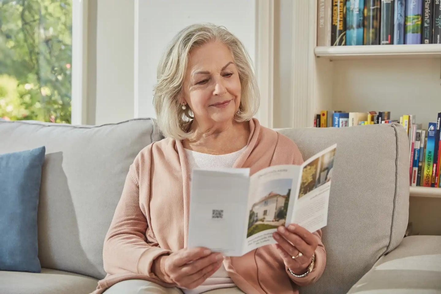 Eine Frau im Rentenalter sitzt auf einem Sofa und liest in einem Prospekt.