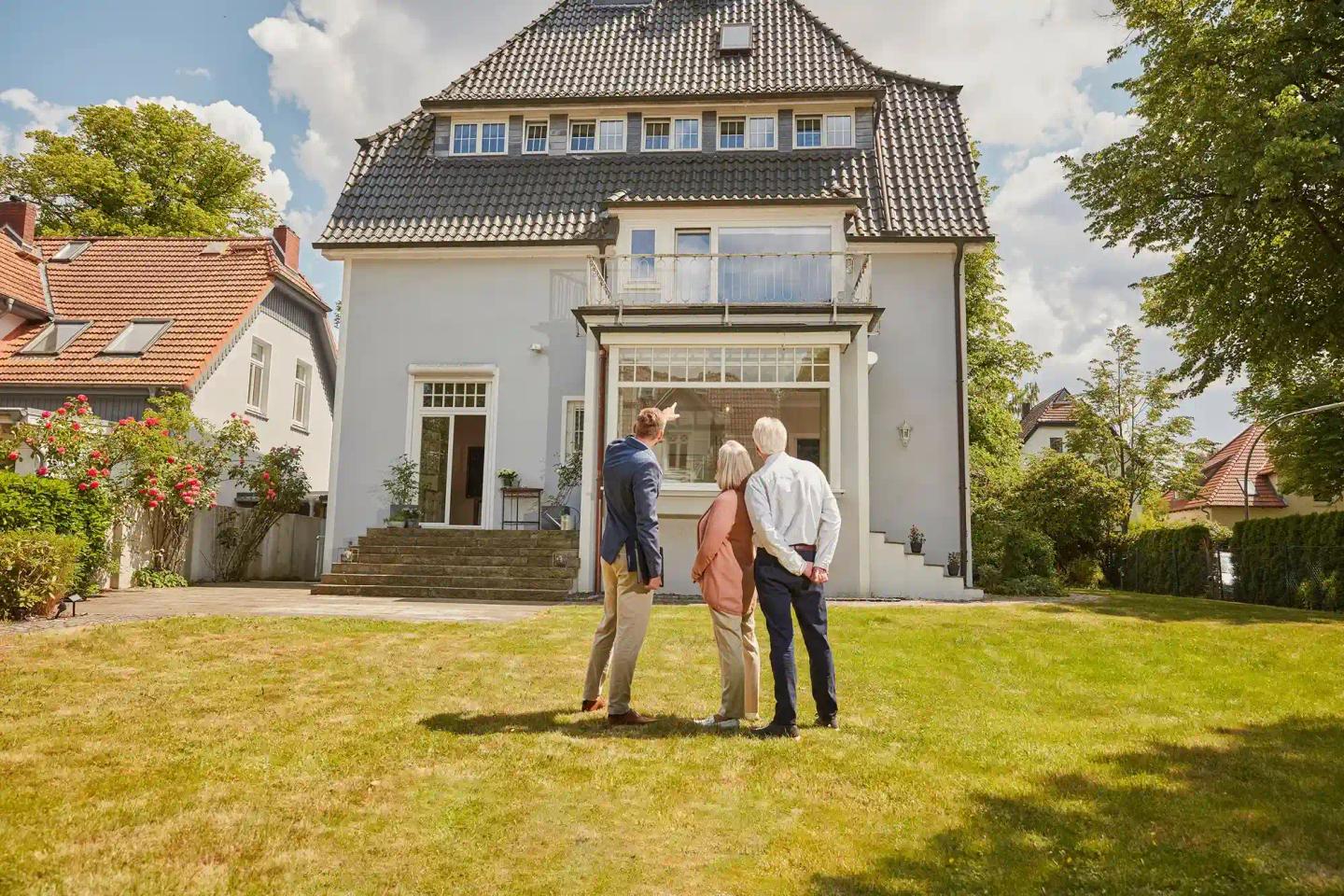 Ein Gutachter im Anzug steht mit einem Ehepaar im Garten, sie schauen gemeinsam auf eine Immobilie. Sie besprechen die Immobilienbewertung ihres Hauses.