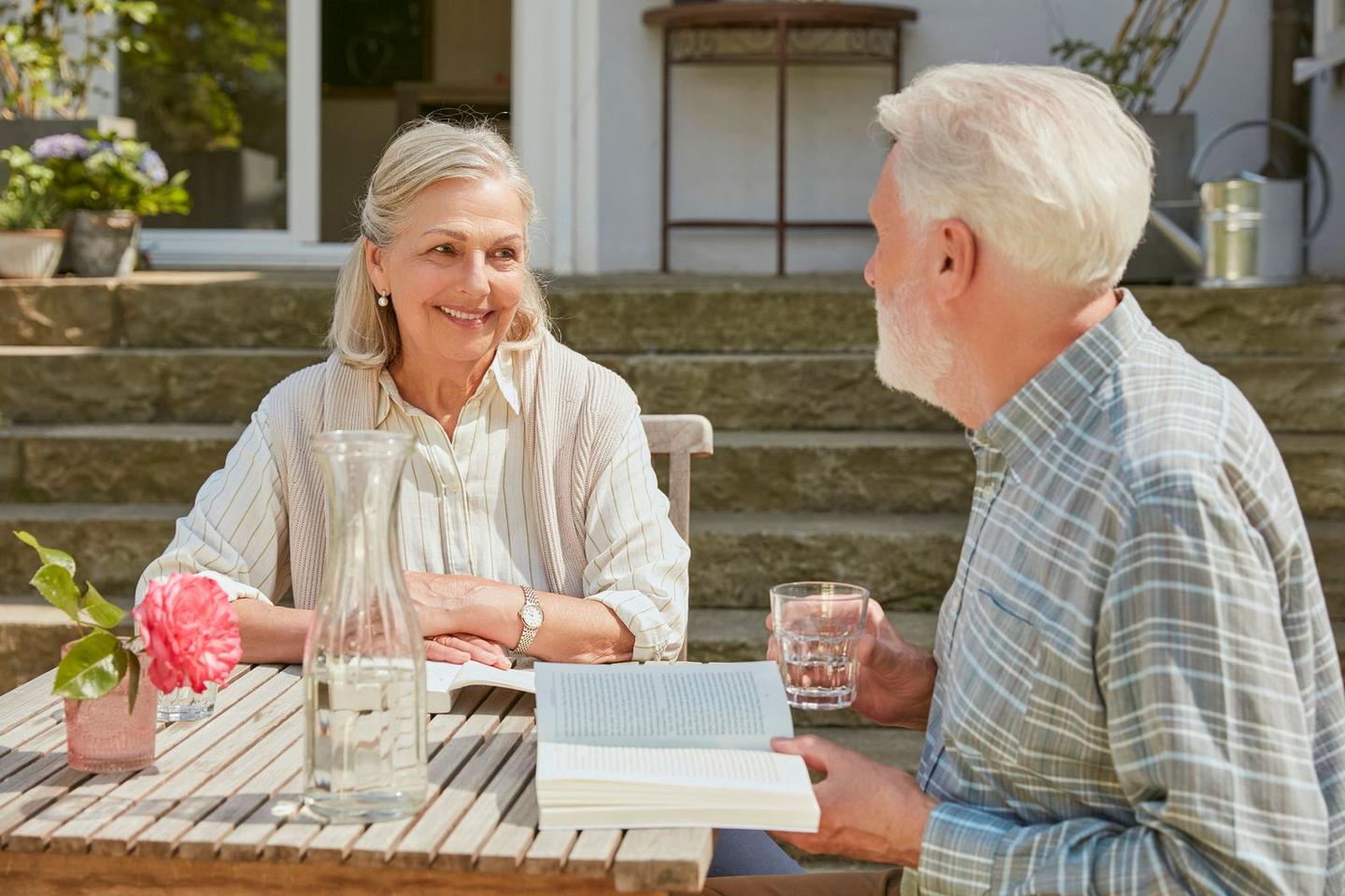 Ein älteres Ehepaar sitzt an einem Gartentisch und trinkt Wasser und lesen ein Buch. Die Frau lächelt ihren Mann an.