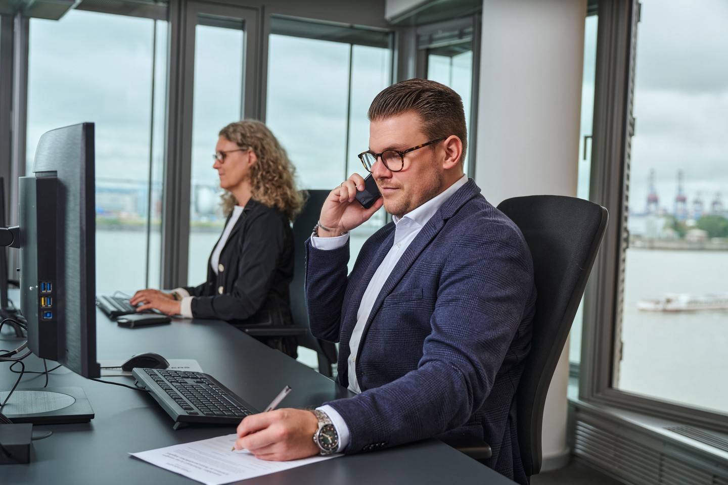 Zwei Kundenberater sitzen nebeneinander. Der Mann ist am Telefon, die Frau arbeitet am Laptop. Sie informieren über den Teilverkauf.