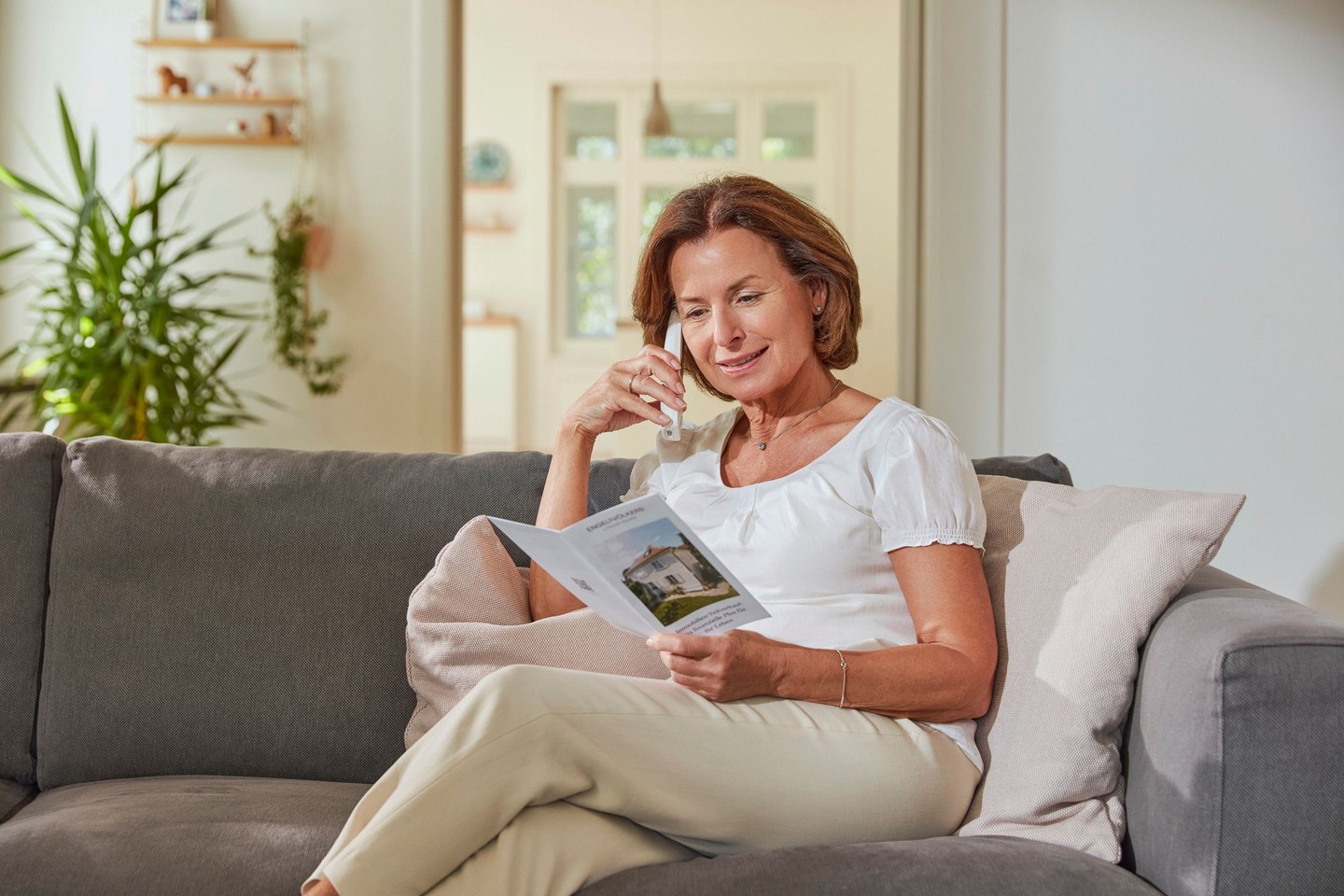 Eine Frau sitzt auf einem Sofa und spricht am Telefon. Sie hält eine Kundenbroschüre über den Teilverkauf in den Händen.