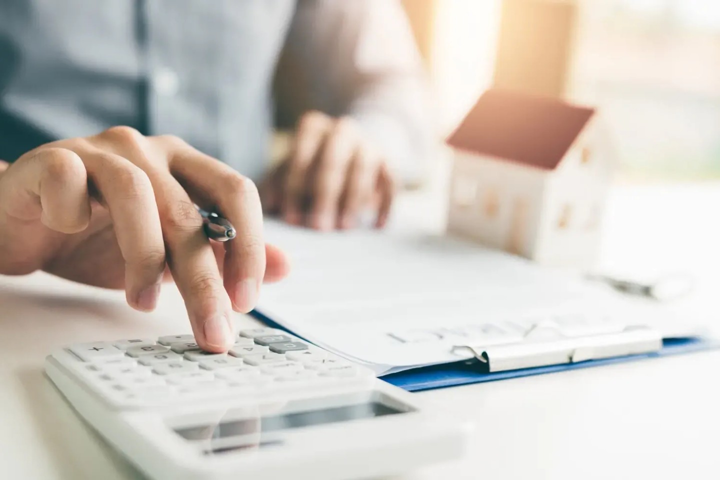 Eine Hand rechnet etwas an einem Taschenrechner. Ein Dokument liegt auf dem Tisch und eine kleine Figur eines Hauses steht daneben. Er rechnet die Steuern die bei einem Immobilienkauf anfallen aus.