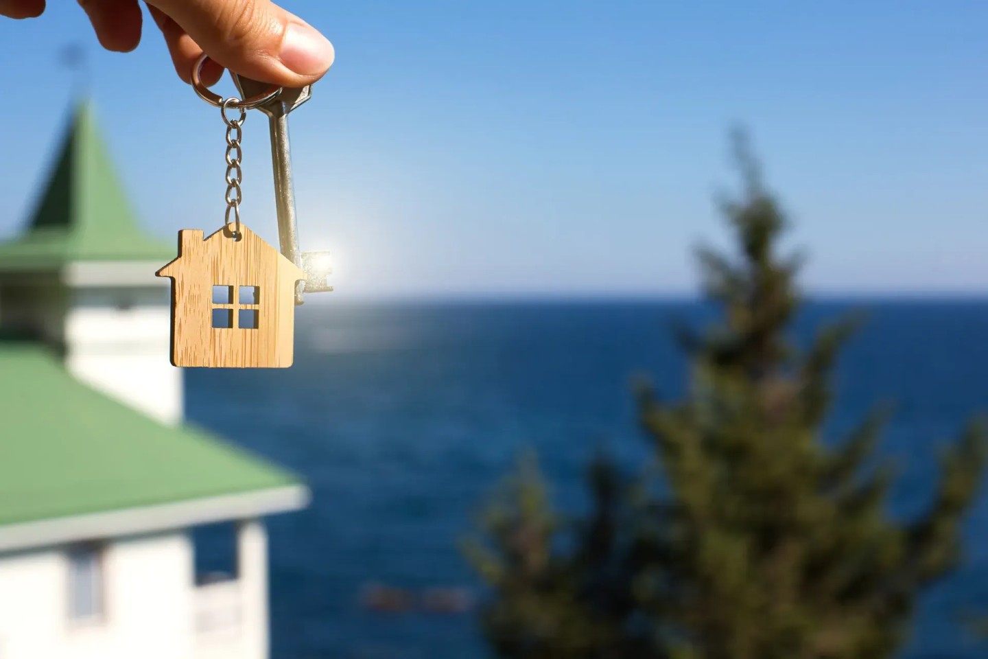 Eine Hand hält einen Schlüssel über einem Haus am Meer. Ein Ferienhaus wurde gekauft mit dem Geld aus dem Teilverkauf.