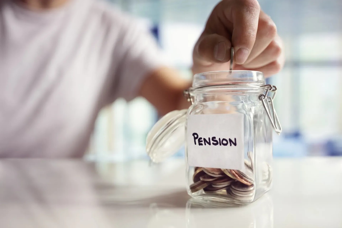 Eine Hand wirft Geldmünzen in ein Glas auf dem das Wort ''Pension'' abgebildet ist. Sie spart für ihre Grundrente.