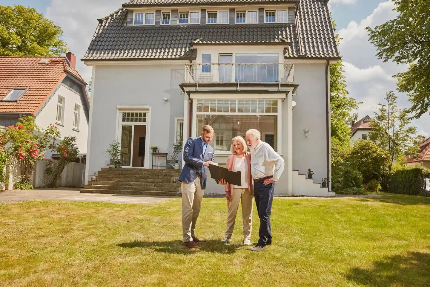 Ein Ehepaar im Rentenalter steht mit einem Mann im Anzug vor einem Haus.