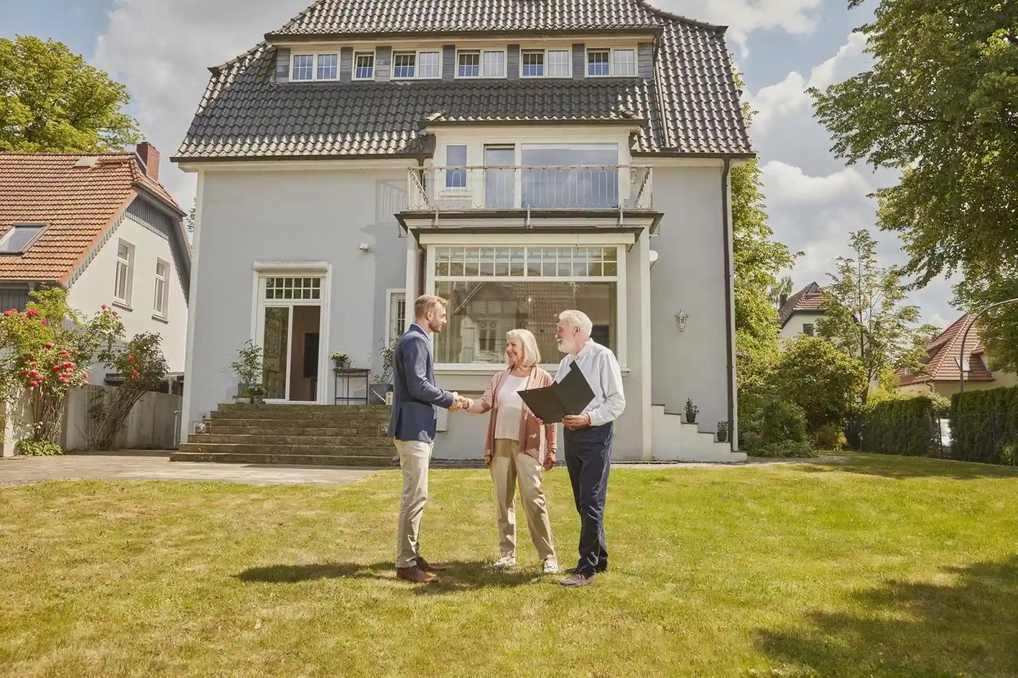 Ehepaar im Rentenalter stehen mit einem Mann im Anzug vor einem Haus auf der Rasenfläche.