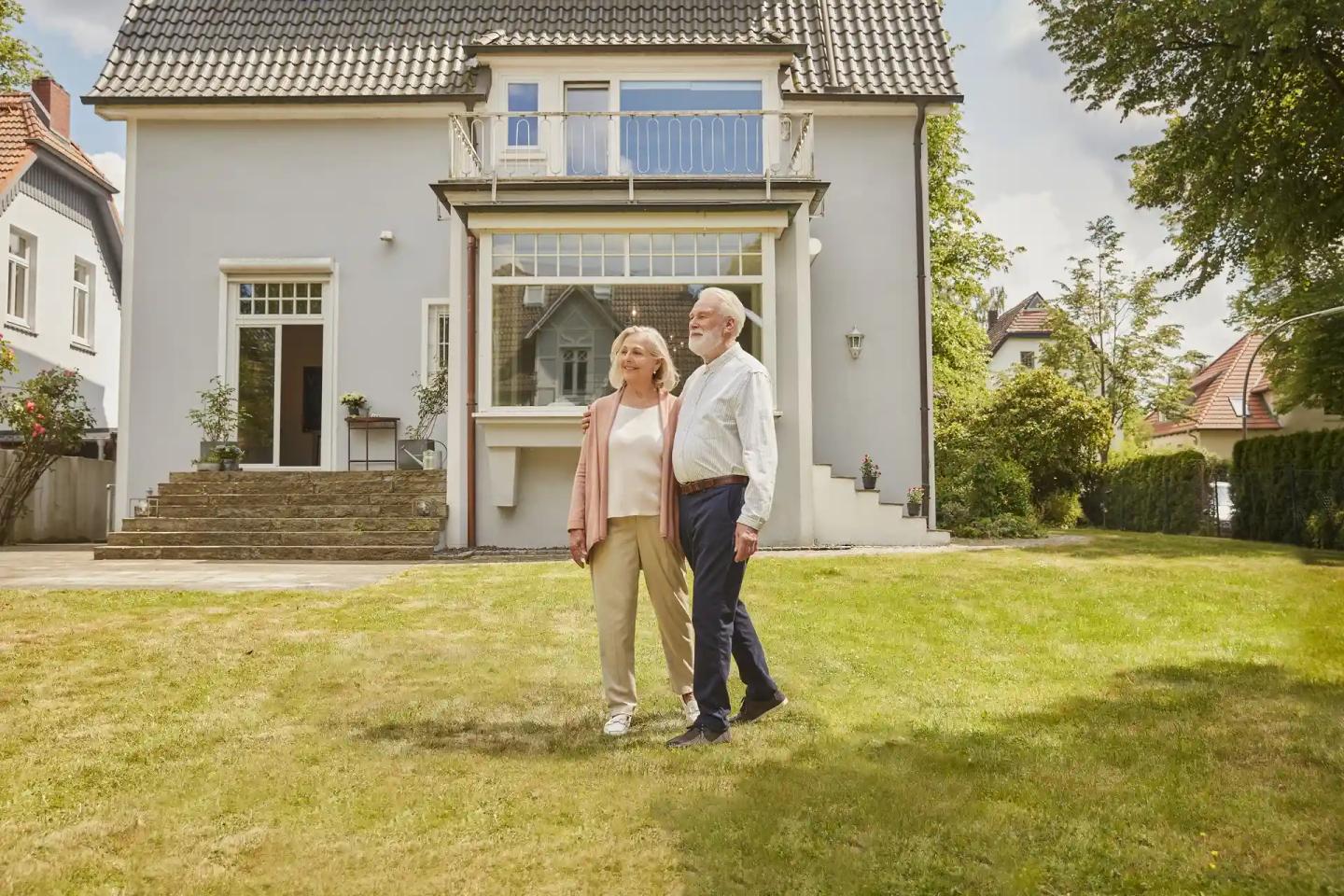 Ein Ehepaar im Rentenalter spaziert durch ihren Garten, im Hintergrund steht ihr Haus.