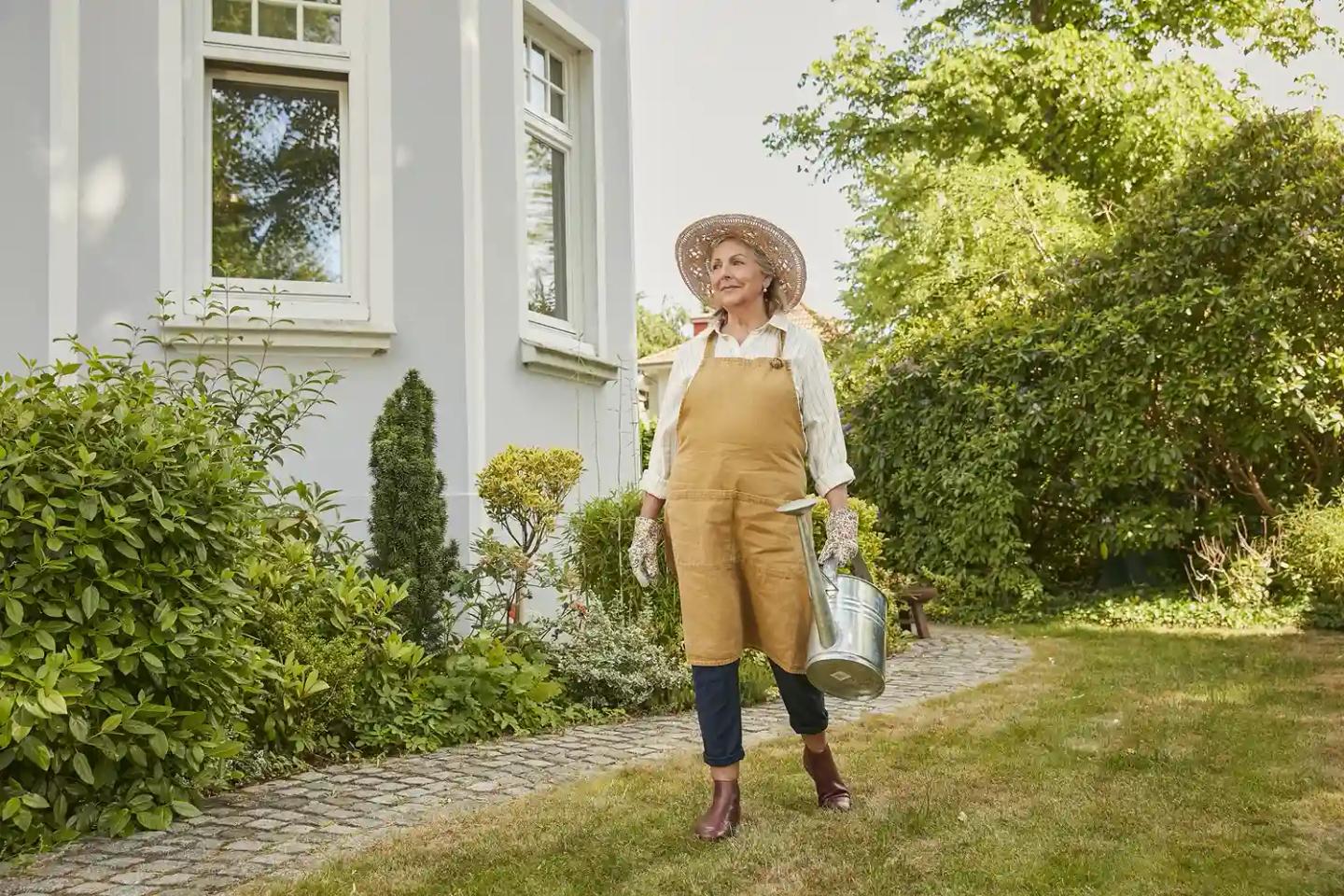 Eine Frau im Rentenalter steht mit einer Gießkanne vor dem Haus im Garten.