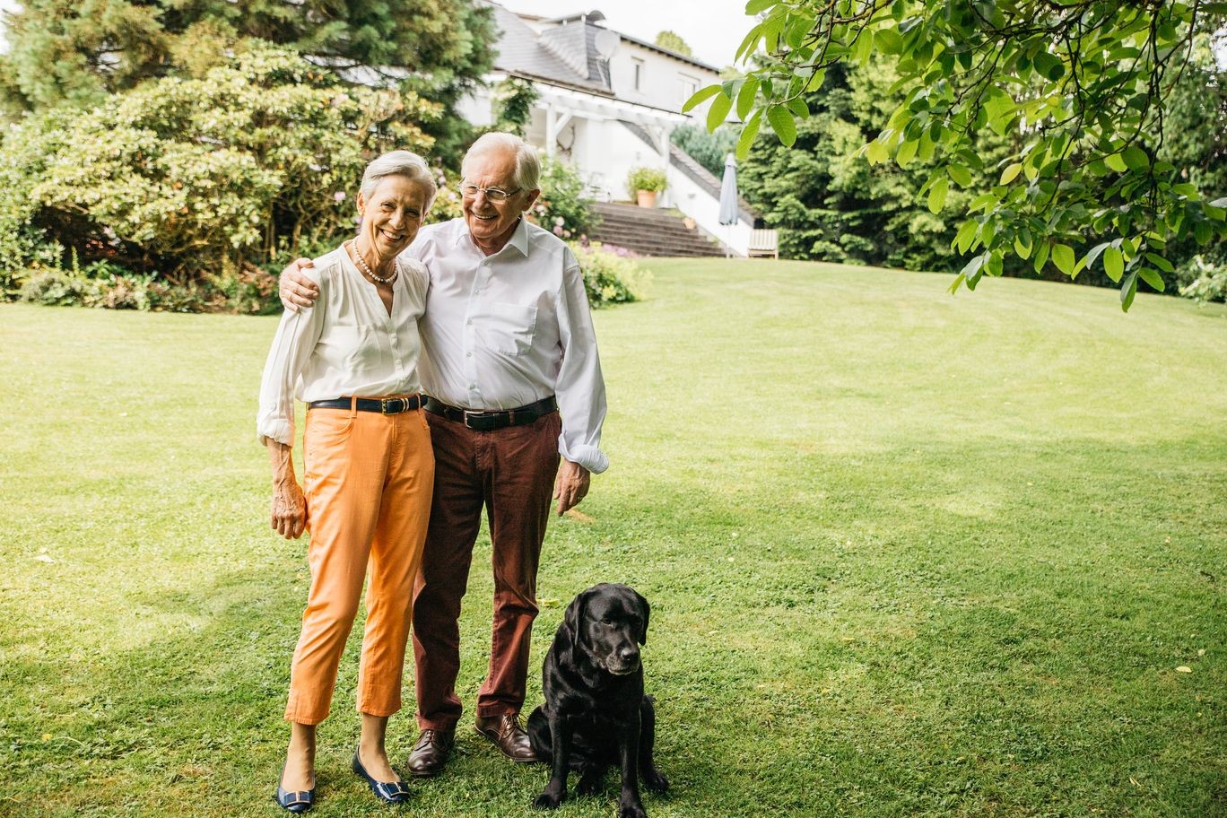 Zwei Kunden stehen mit ihrem schwarzen Labrador vor ihrem Haus im Garten.