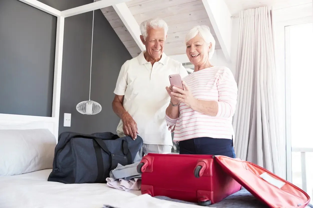 Ein Ehepaar packt ihre Koffer. Sie schauen sich auf dem Handy Destinationen für Seniorenreisen an.
