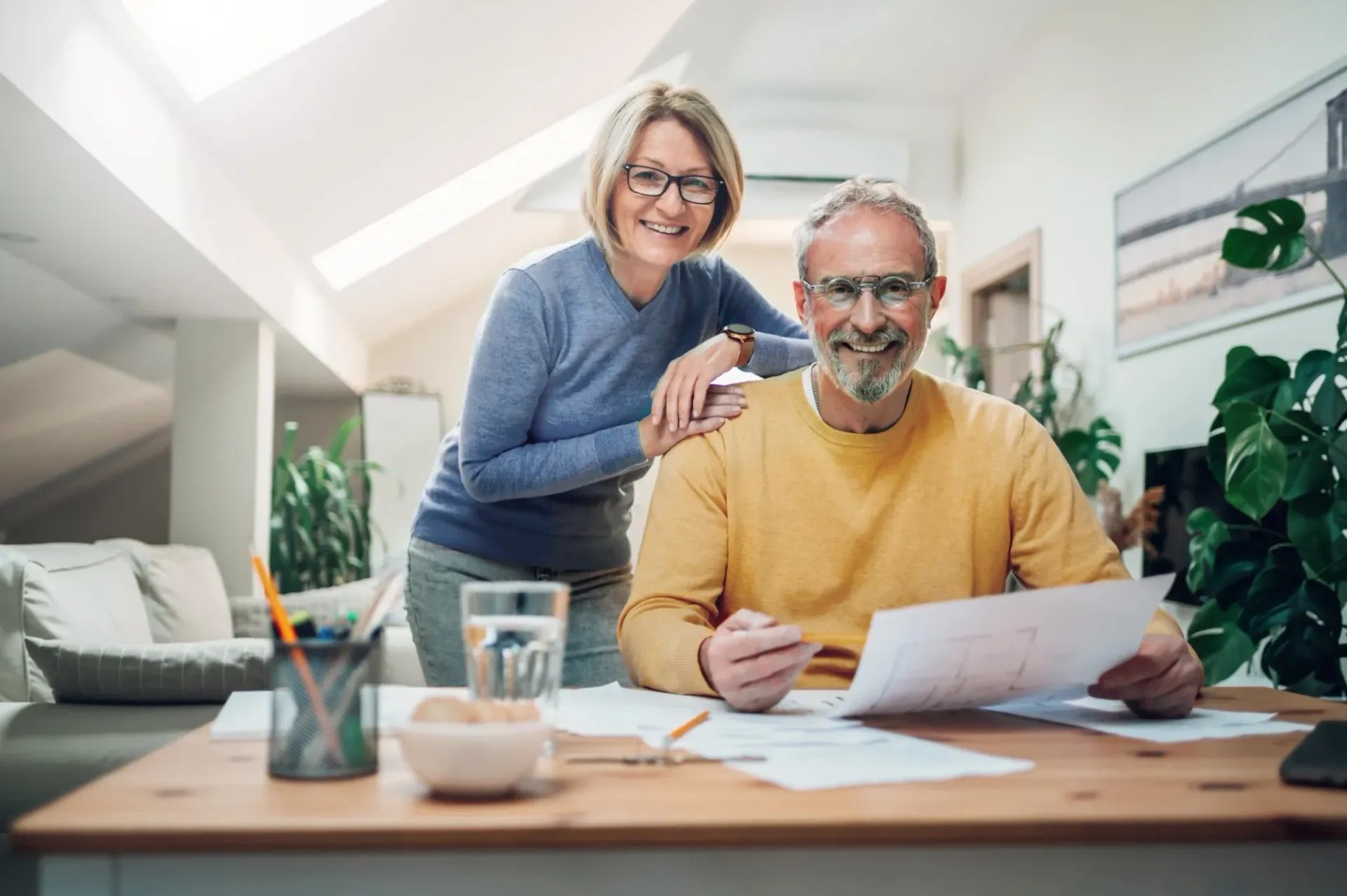 Eine Frau lehnt sich über die Schulter ihres Mannes. Sie schauen sich Dokumente an und überlegen sich die Geldanlage.