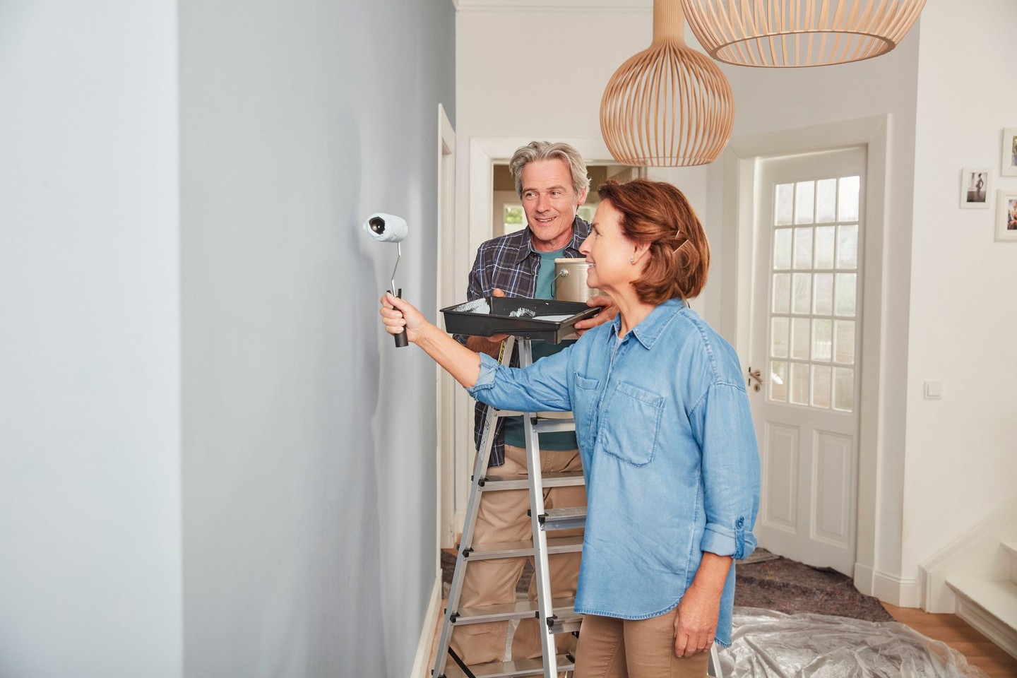 Eine Frau streicht eine Wand in einer grauen Farbe. Ihr Mann steht hinter ihr an einer Leiter und hält die Farbe.