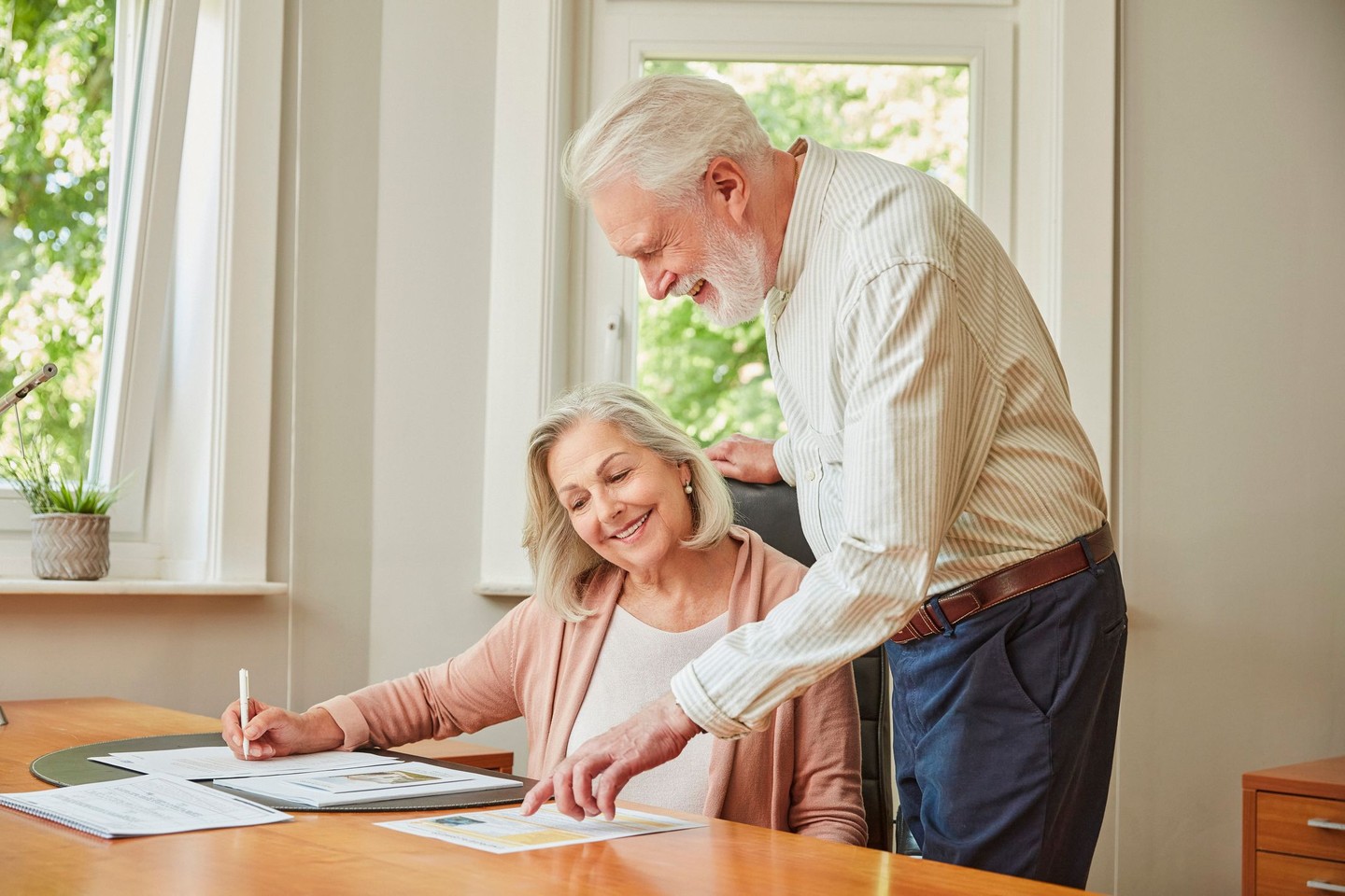 Eine Frau sitzt am Schreibtisch. Ihr Mann ist über sie gebeugt und ließt in einem Dokument.