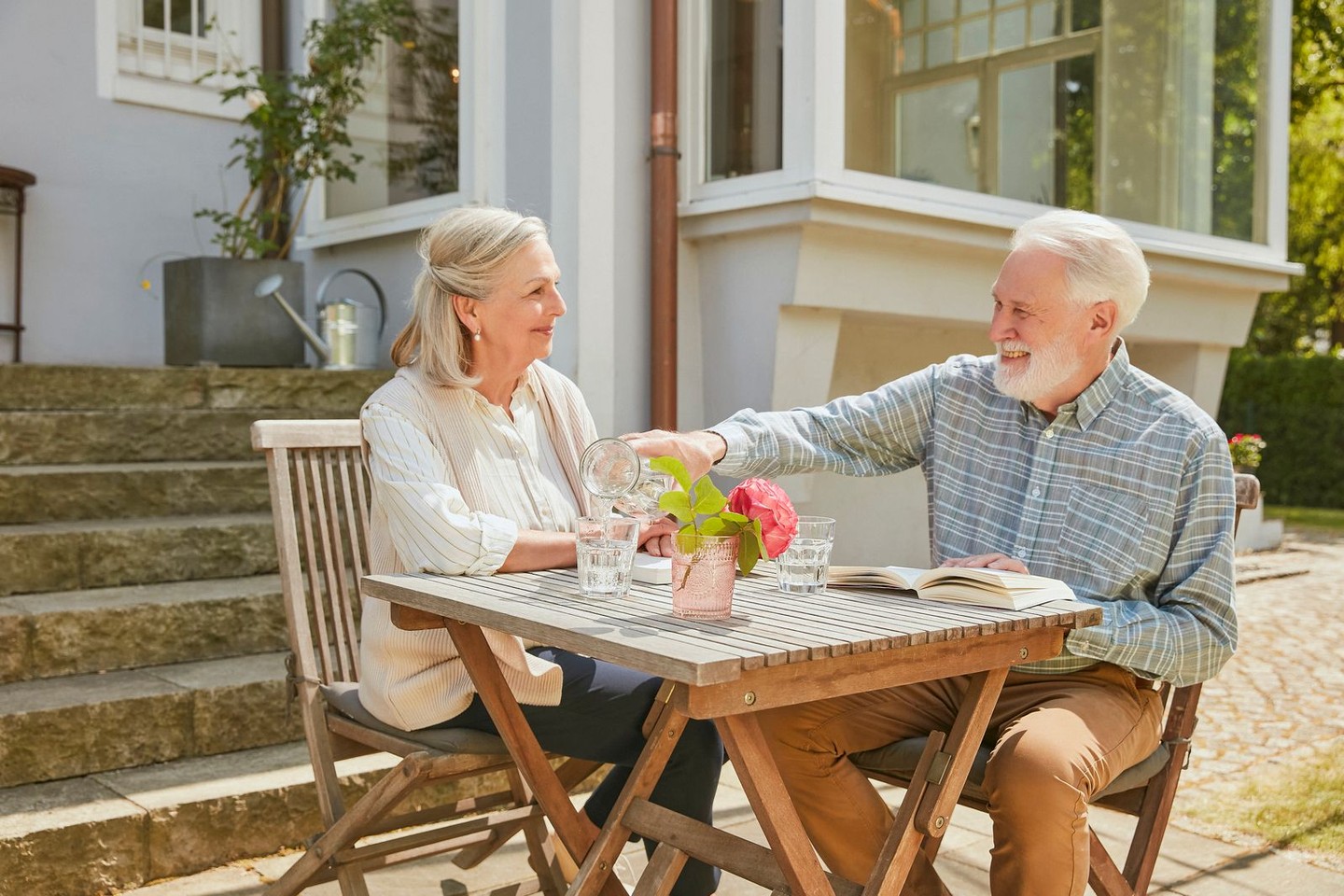 Ein Ehepaar sitzt an einem Gartentisch und trinkt Wasser. Sie lesen ein Buch.
