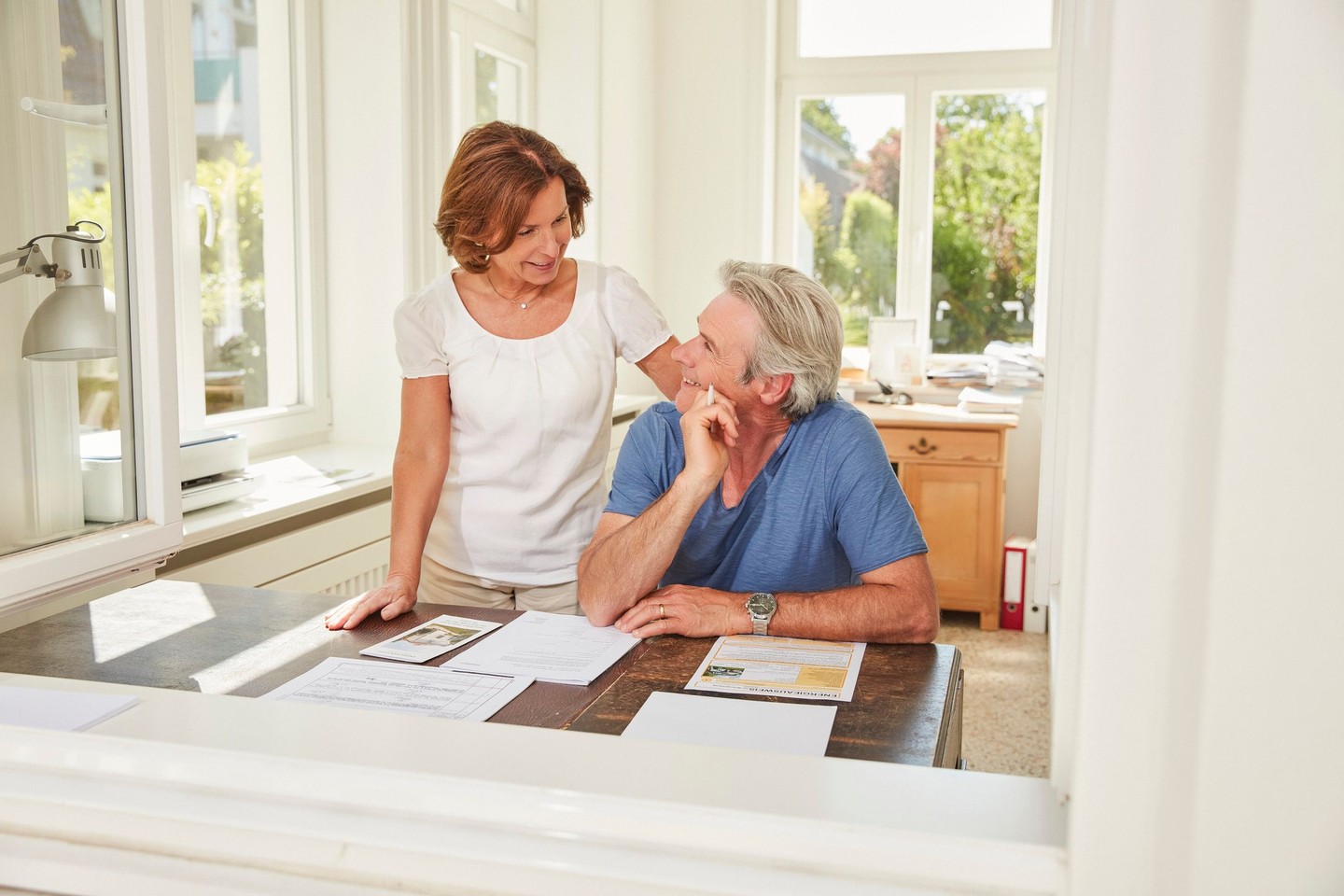 Ein Mann sitzt am Schreibtisch. Seine Frau ist über ihn gebeugt. Auf dem Tisch liegen Dokumente.