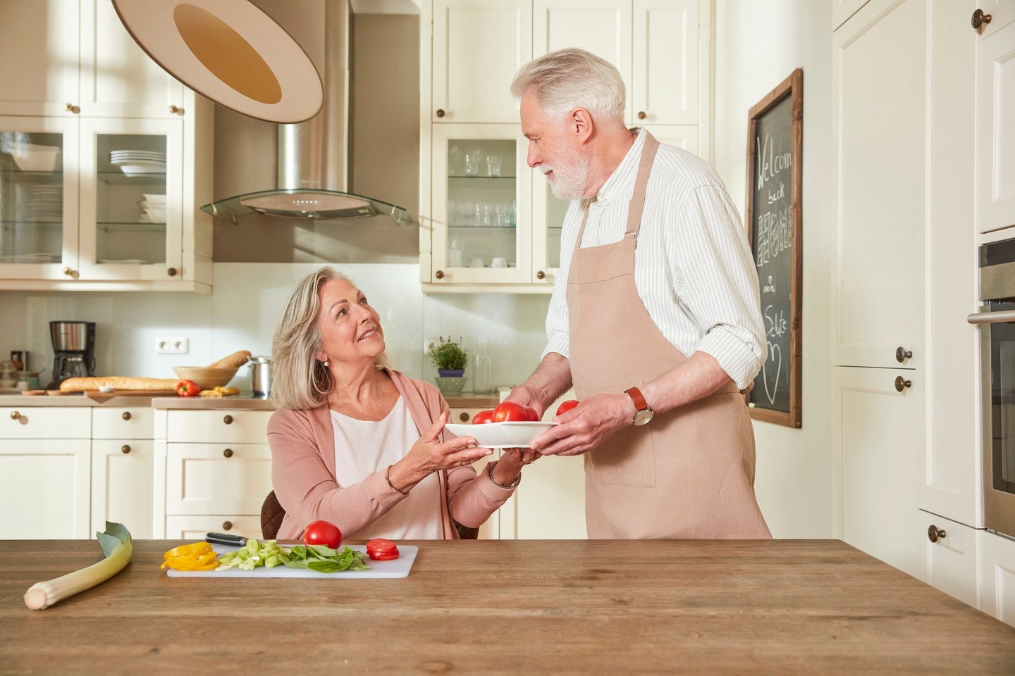 Eine Frau sitzt am Küchentisch. Sie gibt ihrem Mann eine Schale mit frisch geschnittenem Gemüse.