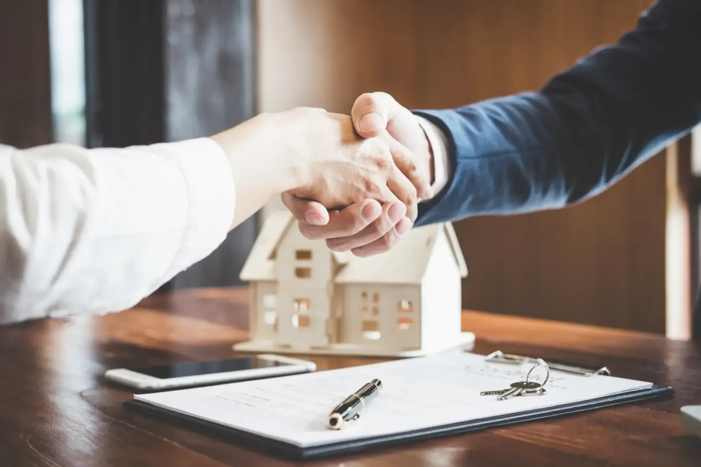 Zwei Personen geben sich die Hand über dem Tisch. Auf dem Tisch liegt ein Papier und eine Figur eines Hauses. Sie haben einen Immobilienkredit abgeschlossen.